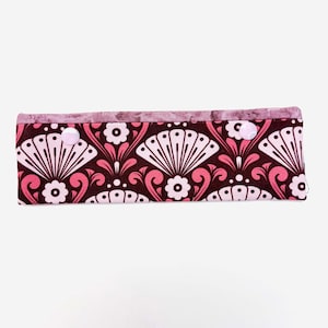 May include: A rectangular fabric case with a repeating pattern of white fan-like shapes, pink flowers, and swirls on a dark brown background. The top edge is a marbled pink fabric, and it fastens with two pink buttons.