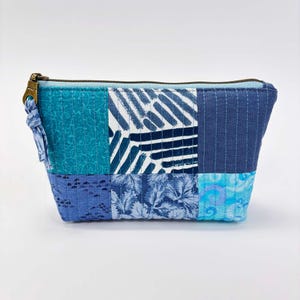 May include: A patchwork zippered pouch in shades of blue and teal. The rectangular bag features a variety of fabric patterns, including swirls, stripes, and floral designs. A brass-coloured zipper and a small fabric loop complete the design.