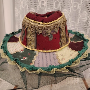 May include: A decorative hat with a patchwork brim and a burgundy crown. The brim features green, yellow, and purple fabric pieces, trimmed with green lace and a black mesh. The crown is adorned with gold trim.