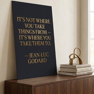Puede incluir: Una impresión artística enmarcada en negro con letras doradas que muestra una cita de Jean-Luc Godard. La impresión está sobre un mueble de madera oscura, junto a una escultura dorada y una pila de libros.