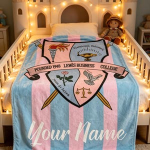 May include: A blue and pink striped blanket with a crest design and the text "Your Name". The crest includes a torch, lamp, rose, dove, and scales. The blanket also has the text "FOUNDED 1948 LEWIS BUSINESS COLLEGE".