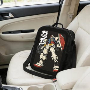 May include: Black lunch bag with a shoulder strap featuring a colorful illustration of a robot with a red shield and the Japanese text "ガンダム". The bag is sitting on a white car seat.