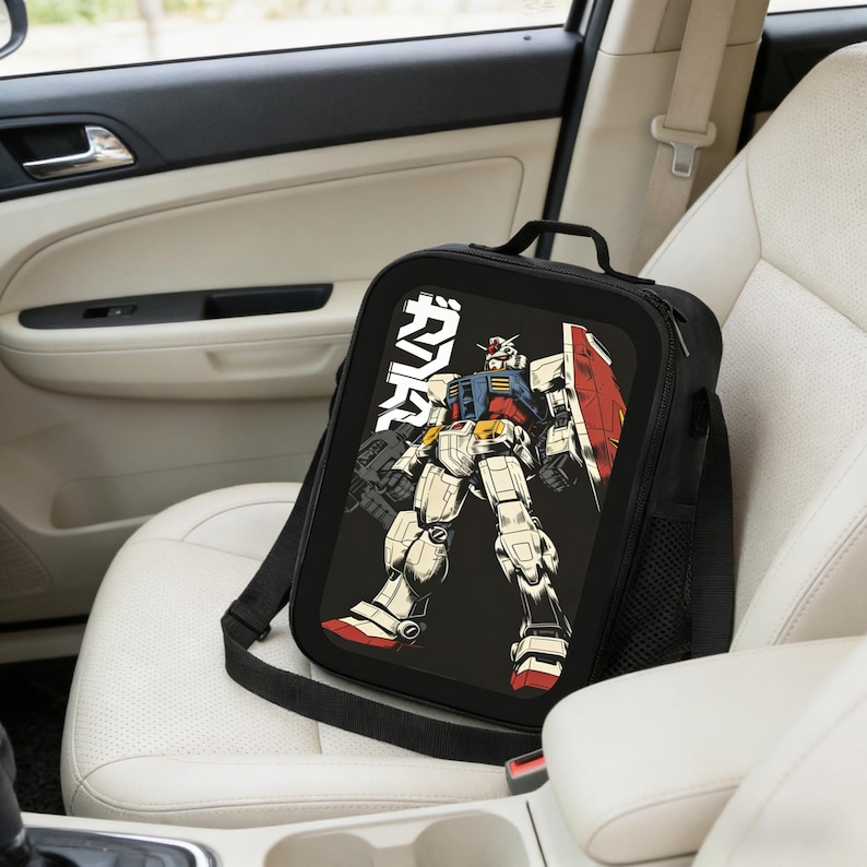 May include: Black lunch bag with a shoulder strap featuring a colorful illustration of a robot with a red shield and the Japanese text "ガンダム". The bag is sitting on a white car seat.