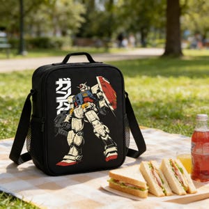 May include: Black lunch bag featuring a colorful illustration of a robot with Japanese text. The bag has a handle, a shoulder strap, and a mesh side pocket. Sandwiches, juice, and a drink are on a checkered blanket.