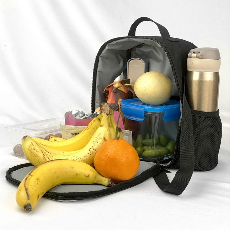 May include: A black insulated lunch bag is open, revealing a variety of food items. Bananas, an orange, and a pear are in front of the bag. Inside, a container with a blue lid, a thermos, and other snacks are visible.