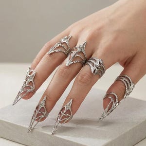 Può includere: Una mano che indossa diversi anelli color argento. Gli anelli includono anelli per unghie lunghi e appuntiti e vari disegni sulle dita. Gli anelli presentano motivi traforati intricati e sono esposti su uno sfondo neutro.