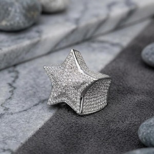 May include: A silver star-shaped ring, encrusted with numerous small, sparkling stones. The ring is set against a gray and white background, with smooth, gray stones scattered around it. The ring's design is bold and eye-catching.