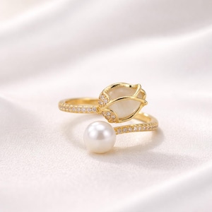 May include: A gold-colored ring with a tulip-shaped design and a pearl. The ring features a band adorned with small, clear stones. The tulip is white with gold accents and small clear stones. The pearl is white and round.