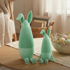 Könnte beinhalten: Zwei mintgrüne, hasenförmige Dekorationsgegenstände stehen auf einem Holztisch. Der größere Hase hat einen hohen, gerippten Körper mit langen Ohren und sitzenden Beinen. Der kleinere Hase hat ein ähnliches Design. Im Hintergrund befindet sich eine Schale mit Ostereiern in Pastellfarben.