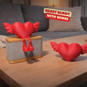 Könnte beinhalten: Zwei rote herzförmige Figuren mit Flügeln. Ein Herz hat Beine und Stiefel und sitzt auf einer grau- und holzfarbenen Box. Die andere Herzfigur befindet sich auf einem Holztisch. Der Text "HEART BUDDY WITH WINGS" befindet sich im Hintergrund.