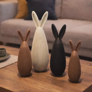 Könnte beinhalten: Vier dekorative Hasenfiguren in Braun-, Weiß- und Schwarztönen. Die Hasen haben lange Ohren und eine gerippte Textur und stehen auf einem Holztisch. Der Hintergrund umfasst ein Sofa mit Kissen.