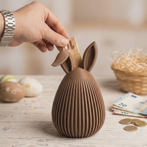 Könnte beinhalten: Ein braunes, eiförmiges Sparschwein mit Hasenohren. Die Spardose hat vertikale Rillen und einen Schlitz zum Einwerfen von Geld. Eine Hand legt einen Geldschein in den Schlitz. Ostereier, ein Korb und Währung sind im Hintergrund.