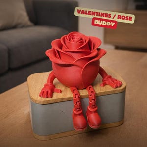 Könnte beinhalten: Rote rosenförmige Figur mit Armen und Beinen, die auf einer grau- und holzfarbenen Box sitzt. Die Figur hat ein lächelndes Gesicht und trägt die Aufschrift "VALENTINES / ROSE BUDDY".