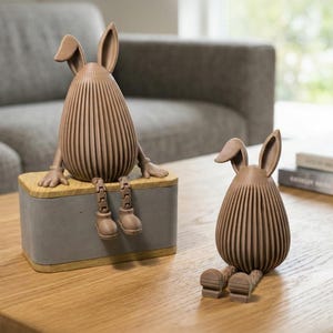 Könnte beinhalten: Zwei braune Hasenfiguren mit vertikal gerippten Körpern und langen Ohren. Einer sitzt auf einer grau- und holzfarbenen Box, die Beine baumeln. Der andere sitzt auf einer Holzoberfläche. Dekorative Osterdekoration.
