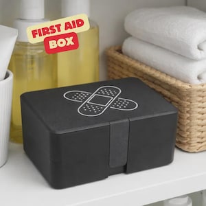 Könnte beinhalten: Eine schwarze Erste-Hilfe-Box mit einem weißen Pflaster-Design auf dem Deckel. Die Box ist rechteckig und steht auf einem weißen Regal. Die Worte "FIRST AID BOX" stehen auf einem gelb-roten Etikett über der Box.