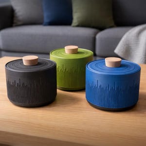 Könnte beinhalten: Drei zylindrische Behälter in Schwarz, Grün und Blau mit Holzdeckeln. Die Behälter haben ein strukturiertes Design um den Sockel. Sie sind auf einem Holztisch angeordnet, mit einem grauen Sofa und Kissen im Hintergrund.