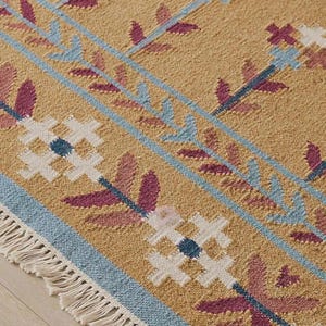 May include: A woven rug with a mustard yellow base featuring a repeating pattern of white cross-shaped flowers, dark pink leaves, and light blue linear borders. The rug is finished with a short, cream-colored fringe along the bottom edge.
