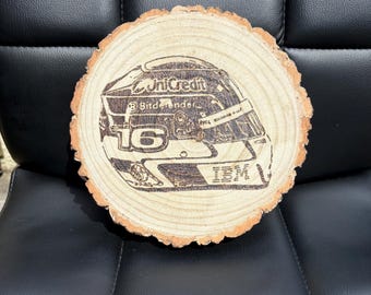 May include: A round wooden coaster with a laser-etched design of a racing helmet. The helmet features the number 16, the UniCredit logo, and the IBM logo. The coaster is made from a natural wood slice with a bark edge.