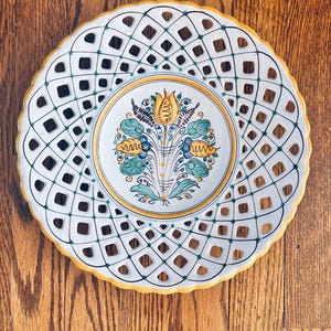 May include: White ceramic decorative plate with a lattice design around the rim. The center features a floral design with yellow, blue, and green accents. The plate has a yellow border and is set against a wooden surface.