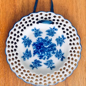 May include: A white porcelain decorative plate with a blue floral design and a scalloped edge. The plate has a series of small holes around the edge and a blue ribbon for hanging. The plate is displayed against a wooden background.