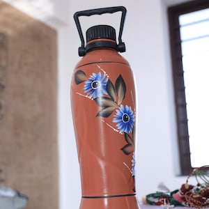 May include: A brown water bottle with a black handle and cap. The bottle is decorated with blue flower designs and dark green leaves. The bottle is sitting on a purple surface.