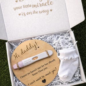 Puede incluir: Una caja de regalo blanca con el texto "Daddy, your little miracle is on the way." En el interior, un disco de madera con "Hi daddy!" y una prueba de embarazo. También se incluye un calcetín de bebé blanco.