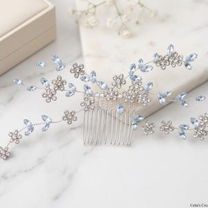 Puede incluir: Un peine de pelo de novia plateado con un diseño floral. El peine presenta cristales transparentes y azul claro dispuestos en un delicado patrón de vid. Los cristales están engarzados en un alambre flexible, creando una apariencia elegante y fluida.