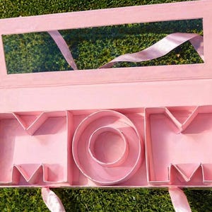 Puede incluir: Una caja de regalo rosa con la palabra "MOM" formada en su interior. La caja tiene una ventana transparente en la tapa y está decorada con cinta rosa. La caja está abierta, revelando los compartimentos interiores que deletrean la palabra.