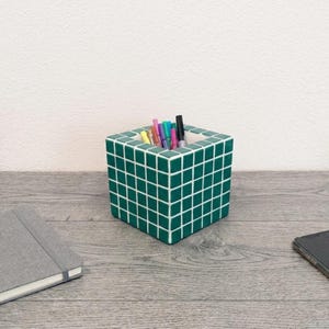 Op de afbeelding: Een vierkante bureau-organizer met een rasterpatroon van kleine, donkergroene tegels met witte voegen. De holle houder is gevuld met diverse gekleurde pennen en stiften, wat een gestructureerde opbergoplossing biedt voor kantoorbenodigdheden.