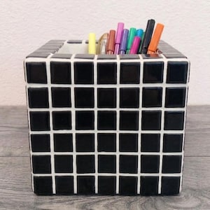 Pode incluir: Um organizador de secretária quadrado com uma grelha de pequenos azulejos pretos brilhantes separados por linhas de argamassa branca. O recipiente está cheio com uma variedade de canetas e marcadores coloridos em tons de amarelo, rosa, azul, preto e laranja.