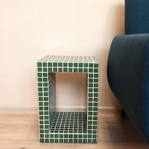 Puede incluir: Una mesa auxiliar rectangular que presenta una cuadrícula de pequeños azulejos cuadrados de color verde oscuro con líneas de lechada blancas. La mesa tiene un diseño abierto y hueco, creando una estructura tipo caja con una superficie plana.