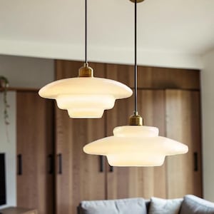 May include: Two pendant lights with a layered, saucer-like design. The lampshades are a soft white, suspended from black cords with brass-colored fixtures. The background includes wooden cabinetry and a gray wall.