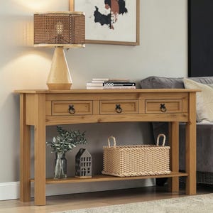 May include: A light-colored wooden console table with three drawers and a lower shelf. A woven basket, a glass vase with greenery, and a decorative house are on the shelf. A lamp with a woven shade sits on the table, and a painting hangs on the wall.