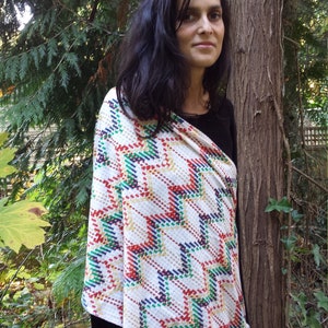 Peut inclure: Un châle blanc avec un motif géométrique coloré en rouge, vert, bleu et jaune. Le châle est drapé sur les épaules, créant un look élégant. L'arrière-plan présente un cadre extérieur naturel avec des arbres.