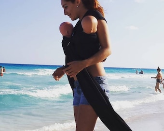 May include: A woman wearing a black baby carrier on a beach. The carrier holds two baby dolls. The woman is wearing denim shorts and is walking near the ocean. The sky is blue and the water is turquoise.
