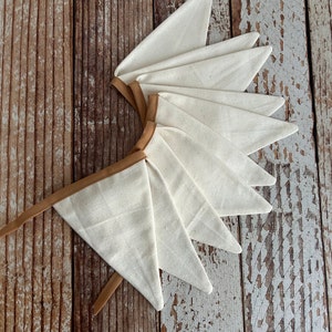 May include: A bunting banner featuring a series of triangular off-white canvas flags attached to a tan fabric ribbon. The flags are arranged in a fan-like overlapping pattern, showcasing the natural texture of the fabric and the clean, straight edges.