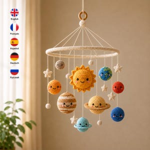 May include: A crochet solar system mobile featuring a central yellow sun surrounded by various planets including Earth, Saturn, and Jupiter, all with embroidered smiling faces. The planets are suspended from a circular frame with white yarn.