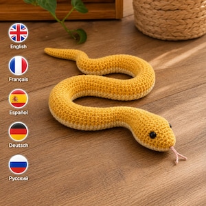 Pode incluir: Um brinquedo de cobra em crochê amarelo com a parte inferior em tom creme mais claro, apresentando dois pequenos olhos de contas pretas e uma língua fina de fio rosa claro saindo da boca. O corpo está enrolado em forma de S.