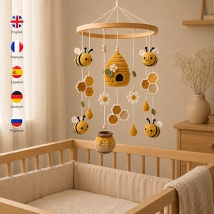 Può includere: Una giostrina per culla all'uncinetto con una cornice circolare in legno da cui pendono api gialle e nere, un alveare giallo centrale, fiori bianchi, forme esagonali a nido d'ape e un piccolo vasetto di miele, sospesi da fili bianchi.