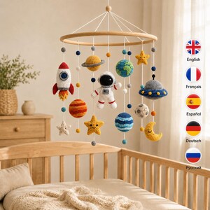 May include: A space-themed nursery mobile featuring crocheted figures of an astronaut, a rocket, planets, stars, and a moon suspended from a circular wooden frame. The colorful yarn elements include a blue and green Earth, a yellow Saturn, and a UFO.