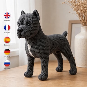 May include: A dark grey crocheted dog figurine standing on four legs. The dog features a textured yarn surface, pointed ears, a short tail, and a distinct white vertical stripe running down the centre of its chest. It has black plastic eyes and a nose.