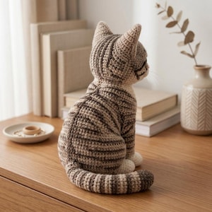May include: A crocheted cat figurine in shades of tan and brown, with dark brown stripes. The cat is sitting upright with its tail curled. The figurine is on a wooden surface, with books and a small vase in the background.