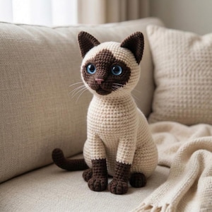 May include: A crocheted Siamese cat doll with a cream-colored body and dark brown accents on the ears, face, paws, and tail. The cat has bright blue eyes and is sitting on a beige sofa. The handmade cat is a decorative item.