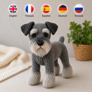 Könnte beinhalten: Eine gehäkelte Schnauzer-Hundefigur mit dunkelgrauem Garn für Körper und Ohren, hellgrauem strukturiertem Garn an den Pfoten und einem markanten weißen Bart. Das Spielzeug hat schwarze Kunststoffaugen und eine schwarze Nase im Stand.