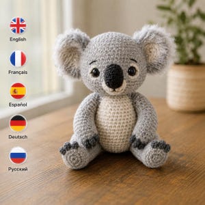 May include: A grey and cream-colored crocheted koala stuffed animal. The koala features a round head with large, fuzzy grey ears, black plastic eyes, and a prominent black nose. The body is grey with a cream-colored belly and dark grey paws.