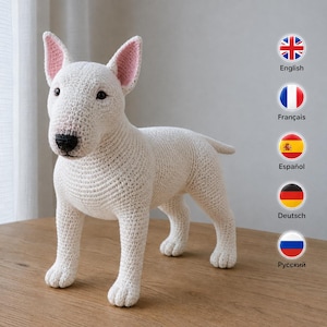 Könnte beinhalten: Eine weiß gehäkelte Bullterrier-Hundefigur mit schwarzer Nase und rosa Innenohren. Das Spielzeug ist in einem strukturierten Maschenmuster gefertigt und verfügt über aufrecht stehende spitze Ohren, dunkle Augen und einen kurzen Schwanz.