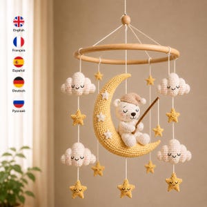 May include: A crochet baby mobile featuring a central yellow crescent moon with a white bear wearing a tan nightcap. The mobile includes white clouds and yellow stars suspended from a circular wooden frame by white strings.