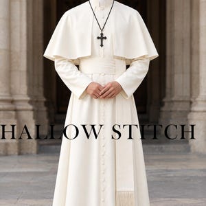 May include: A floor-length off-white cassock featuring a button-down front, a matching wide sash with a fringed end, and a short capelet over the shoulders. A black cord necklace with a dark cross pendant is worn over the high-collared garment.