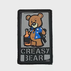 Può includere: Una toppa rettangolare con un orso dei cartoni animati con una camicia blu con accenti di stelle bianche, che tiene in mano una bottiglia e una pistola. Il testo "CREASY BEAR" è visualizzato sotto l'orso in lettere bianche su sfondo nero.