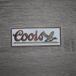 Könnte beinhalten: Gestickter Aufnäher mit dem Wort "Coors" in brauner Schrift und einer fliegenden Entenillustration. Der rechteckige Aufnäher hat einen weißen Hintergrund und einen braunen Rand, für Kleidung oder Accessoires.